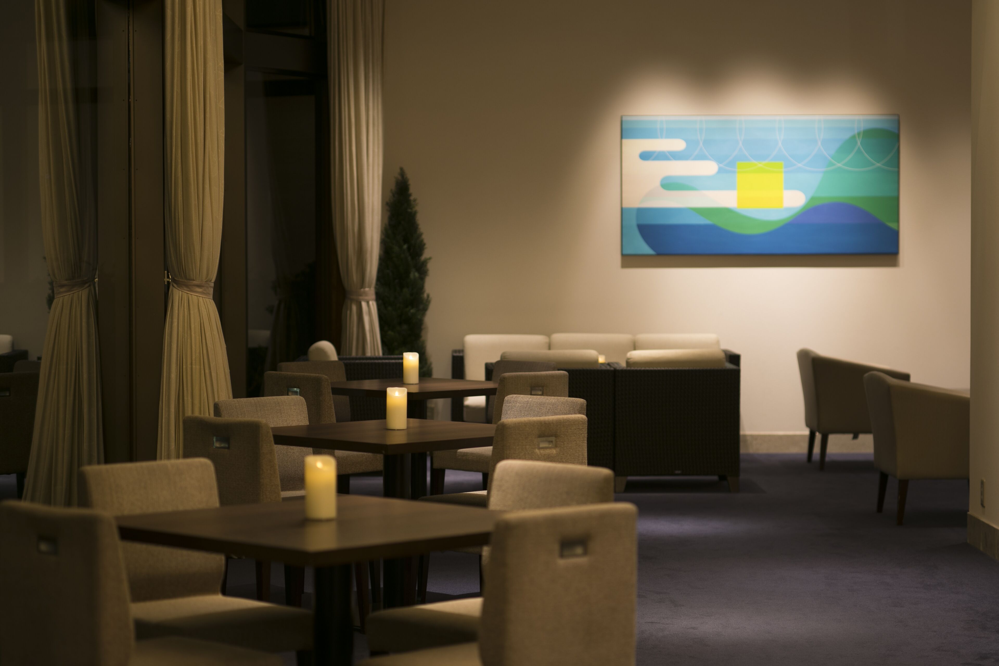A dimly lit room with a sofa, chairs, and tables arranged for dining or socializing. A painting is on the wall, and a candle is on one of the tables.