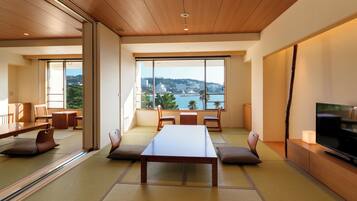 A modern room with tatami mats, a wooden table, chairs, a TV, and a large window with a view of a city and hills.