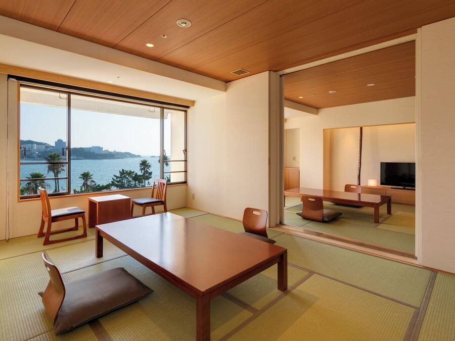 Une chambre moderne dotée d’une grande fenêtre donnant sur la mer et le paysage urbain. Elle comprend une table en bois, des chaises et un téléviseur.