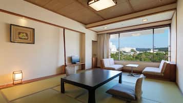 A traditional Japanese room with tatami flooring, a low table, cushioned seating, a television, and a large window offering a city view.
