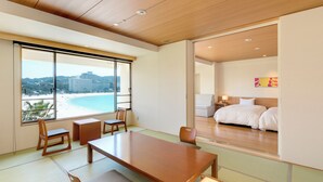 A hotel room with a large wooden table, tatami mats, a bed with white linens, and a view of a beach and mountains through large windows. - Shiraraso Grand Hotel (Shirahama)