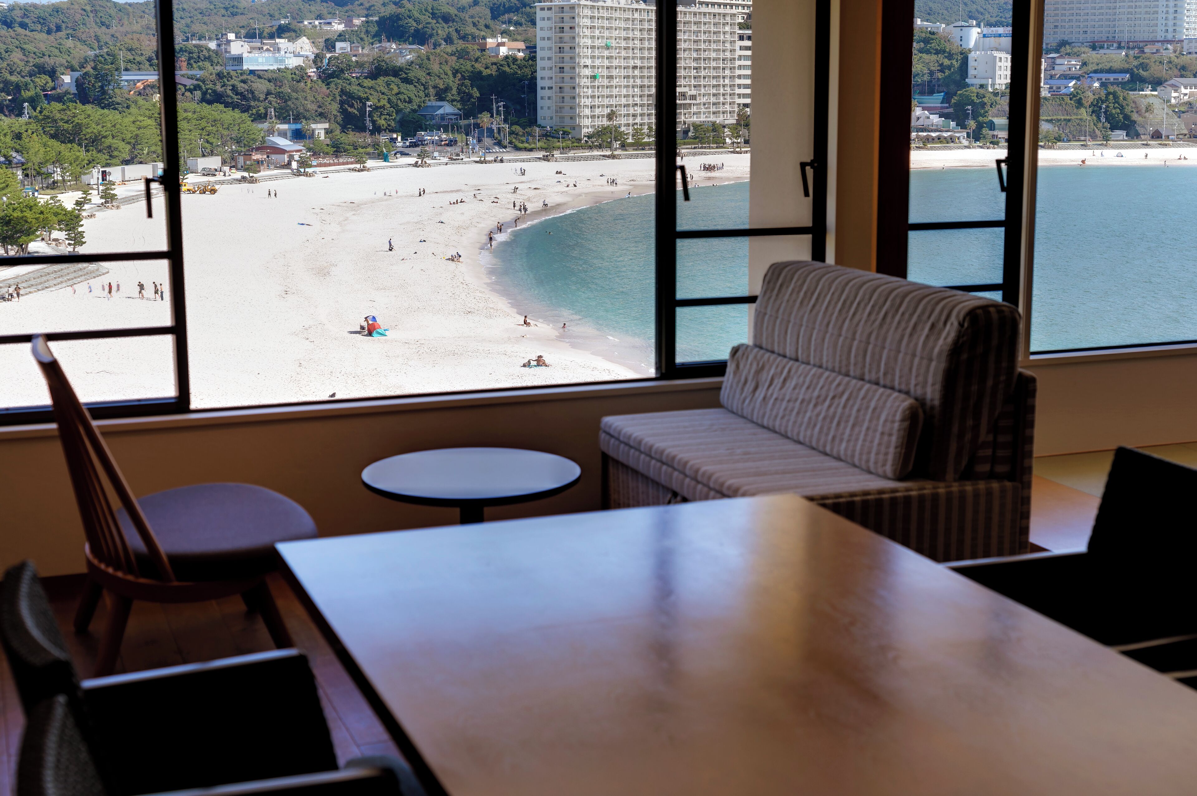 Una habitació amb un finestral gran que dona a una platja, un sofà, una taula petita i una cadira.