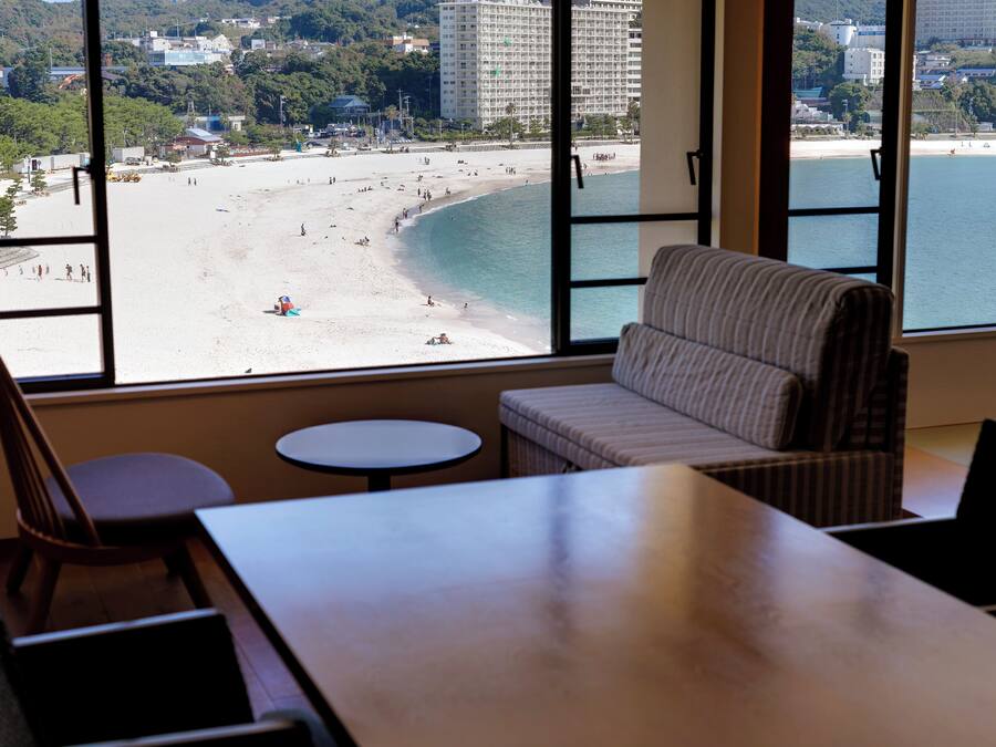 Una habitació amb un finestral gran que dona a una platja, un sofà, una taula petita i una cadira.