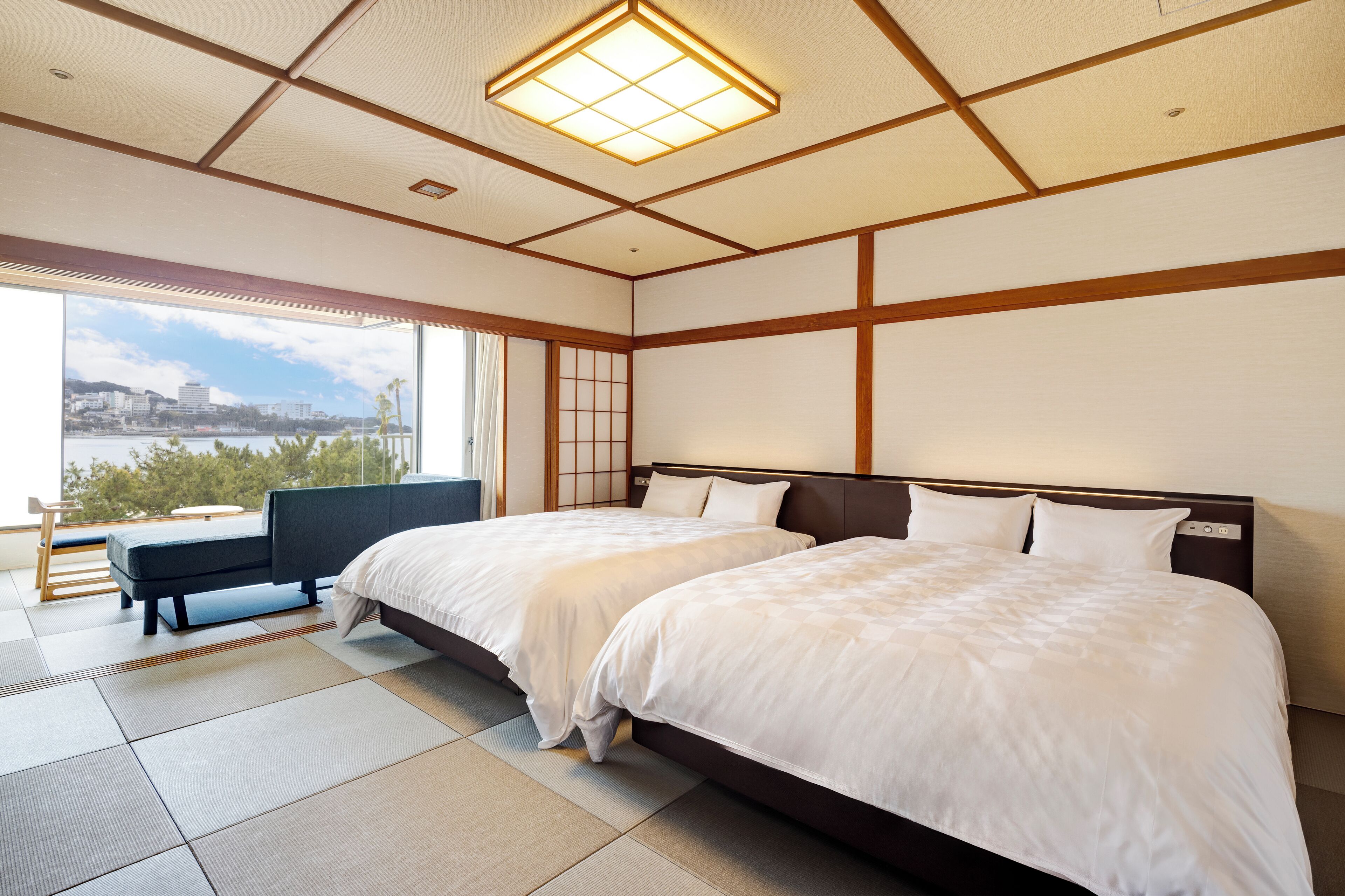 A hotel room with two beds, a bench, a view of a body of water, and a cityscape.