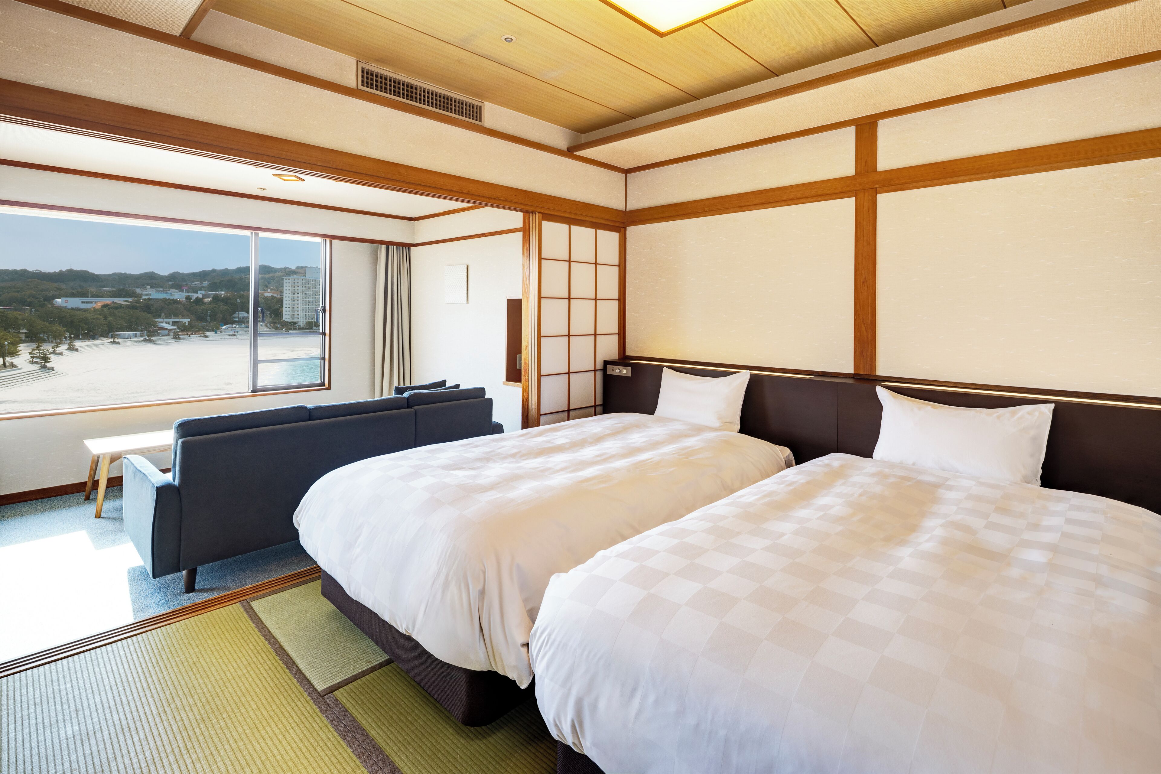 Habitación de hotel con dos camas, un sofá, vista a la playa y paredes de estilo japonés tradicional.