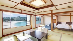 A hotel room with a large window offering a view of a beach and a cityscape. There is a bed, a small table, and a seating area with cushions. - Shiraraso Grand Hotel (Shirahama)