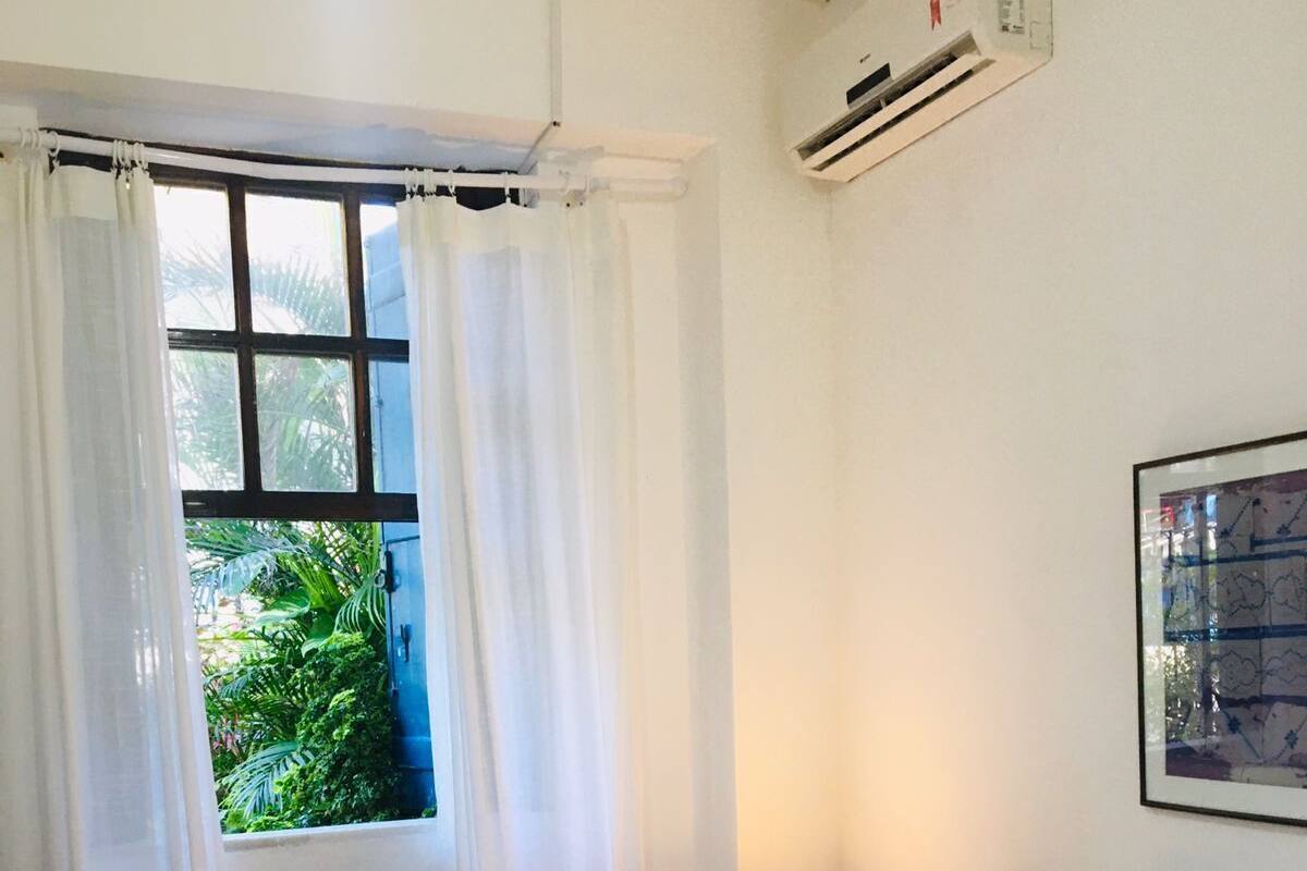 Ein Schlafzimmer mit Bett, weißen Vorhängen, einem Fenster mit Blick auf Grünflächen, einer Klimaanlage und einer Wandlampe.