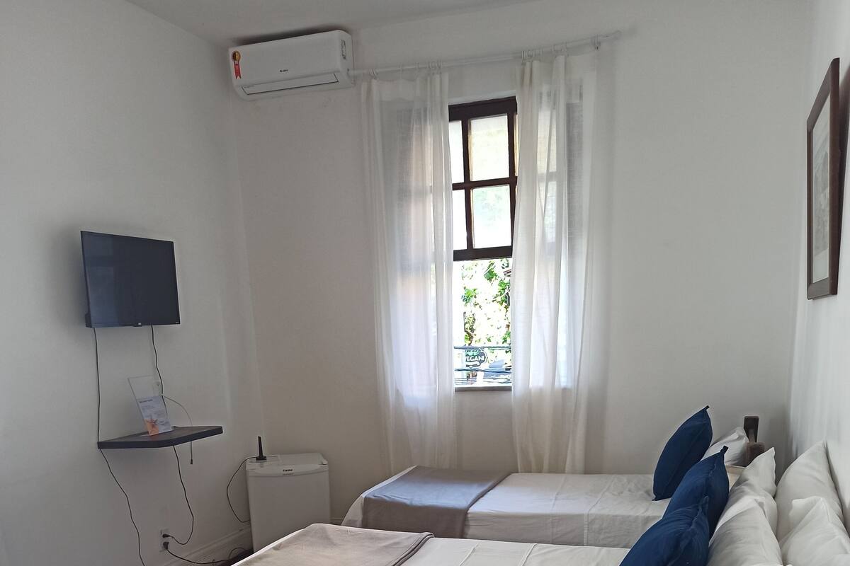 Ein Hotelzimmer mit zwei Betten, einem an der Wand montierten Fernseher, einem Fenster mit Vorhängen und einer Rezeption.