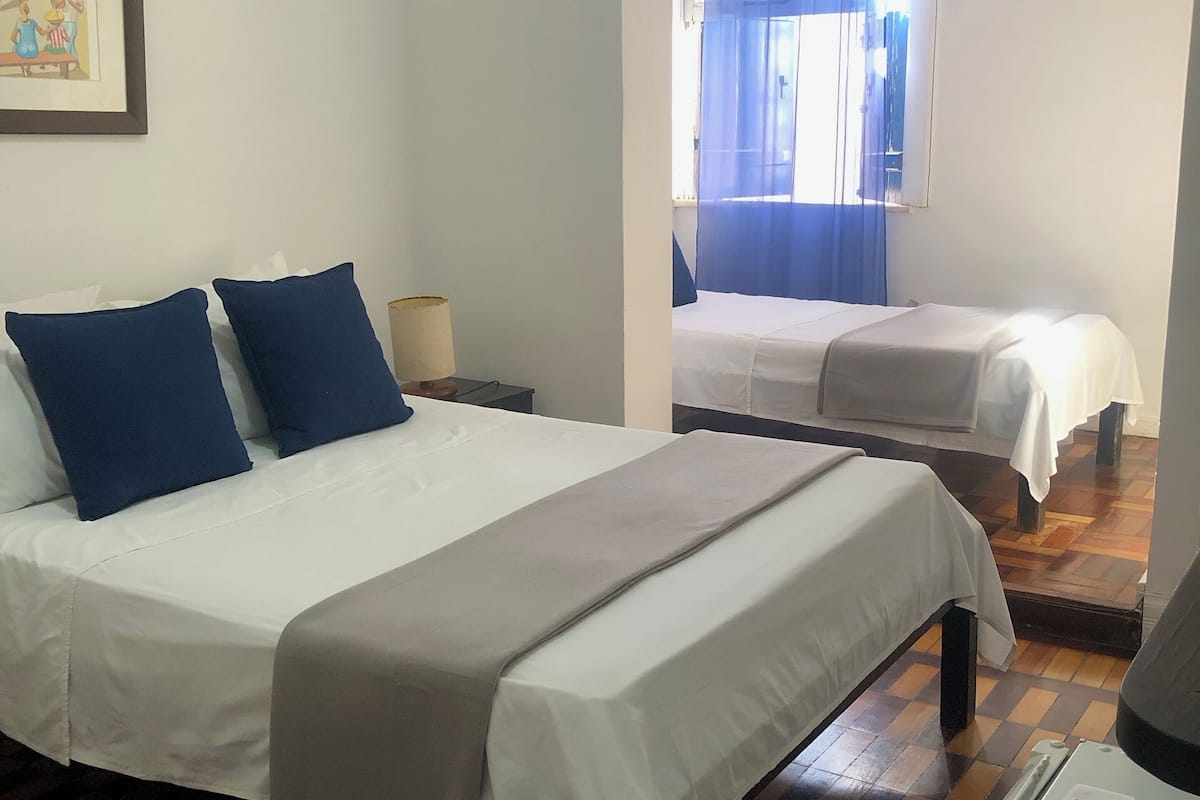 Ein Hotelzimmer mit einem großen Bett, zwei kleineren Betten, einem Schachbrettmuster Boden und einem Fenster mit blauen Vorhängen.