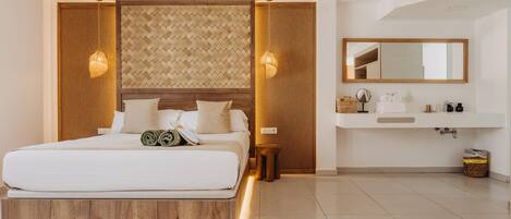 Una habitación de hotel moderna con cabecera de madera, una cama grande, lámparas de noche, y un baño con espejo, lavamanos y artículos de aseo.