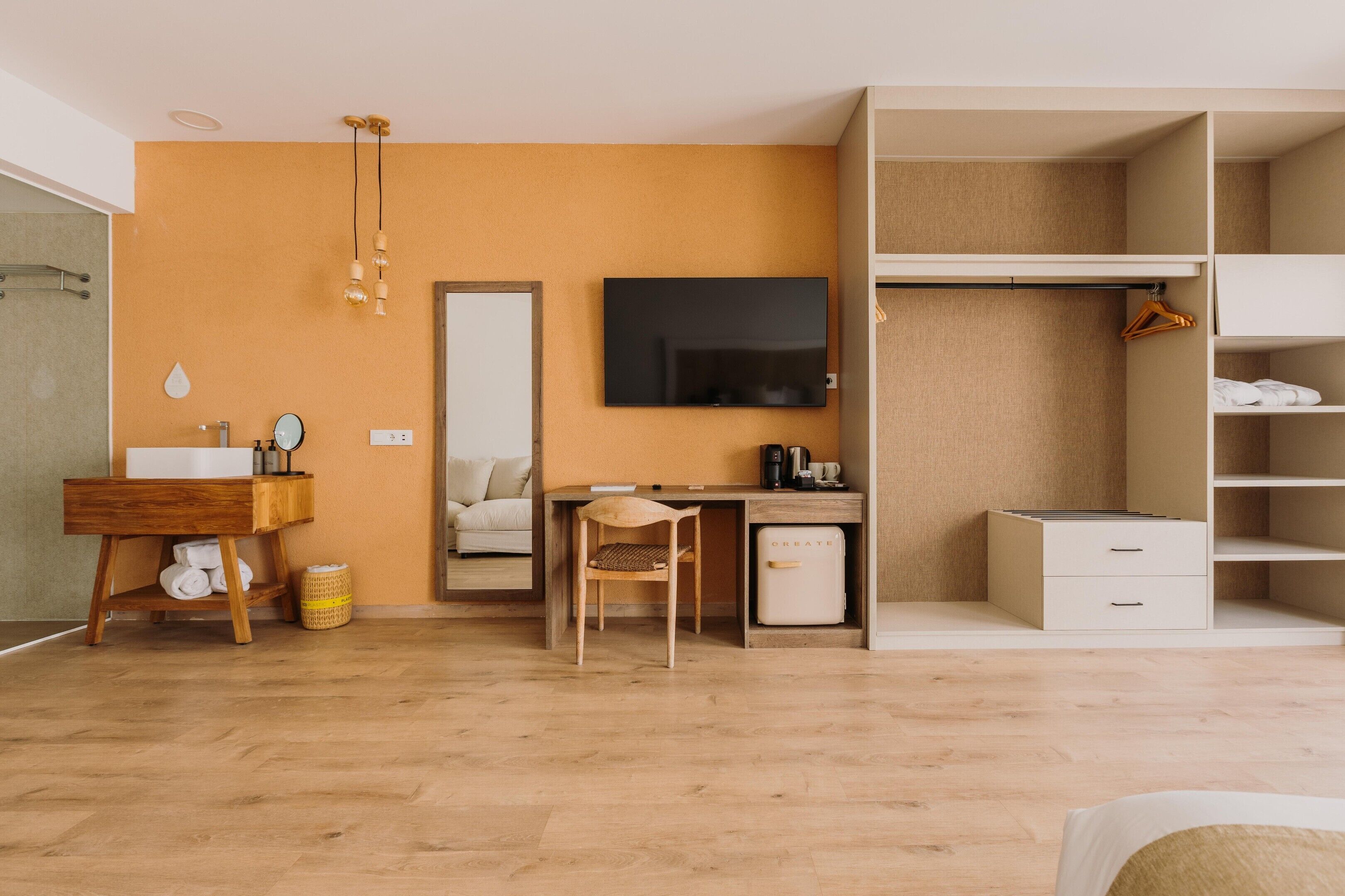 A modern hotel room with a wooden floor, a desk with a chair, a large mirror, a wardrobe, and a television.
