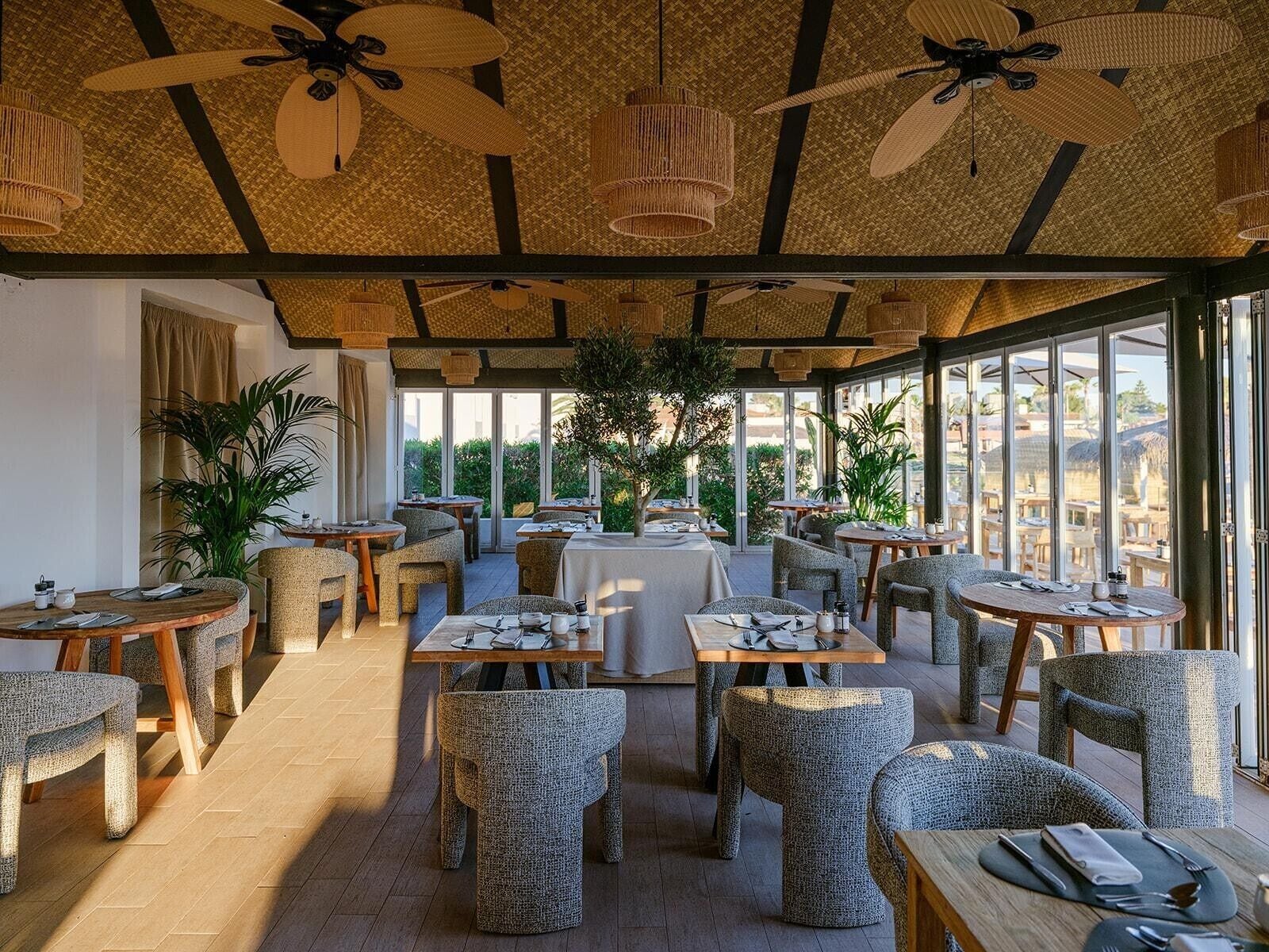 Restaurante com mesas e cadeiras redondas, ventiladores de teto e grandes janelas.