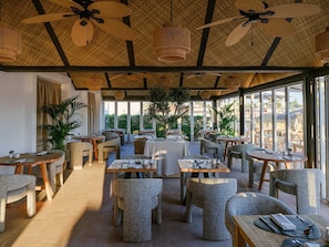 A restaurant with round tables and chairs, ceiling fans, and large windows. - Lago Resort Menorca Casas del Lago - Adults Only (Ciutadella de Menorca)
