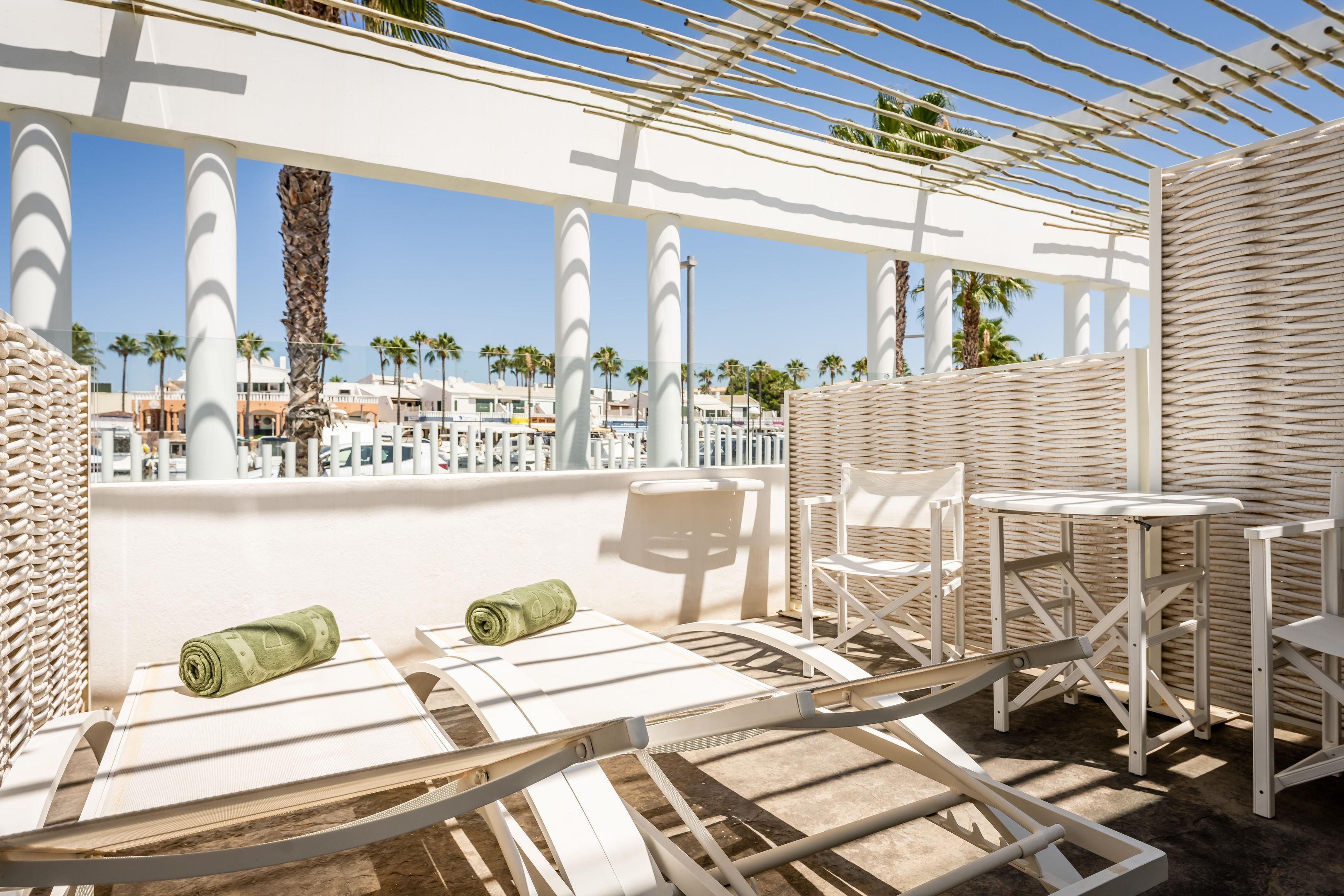 A sunlit outdoor seating area with white chairs and tables, a woven wall, and a view of palm trees and buildings in the background.