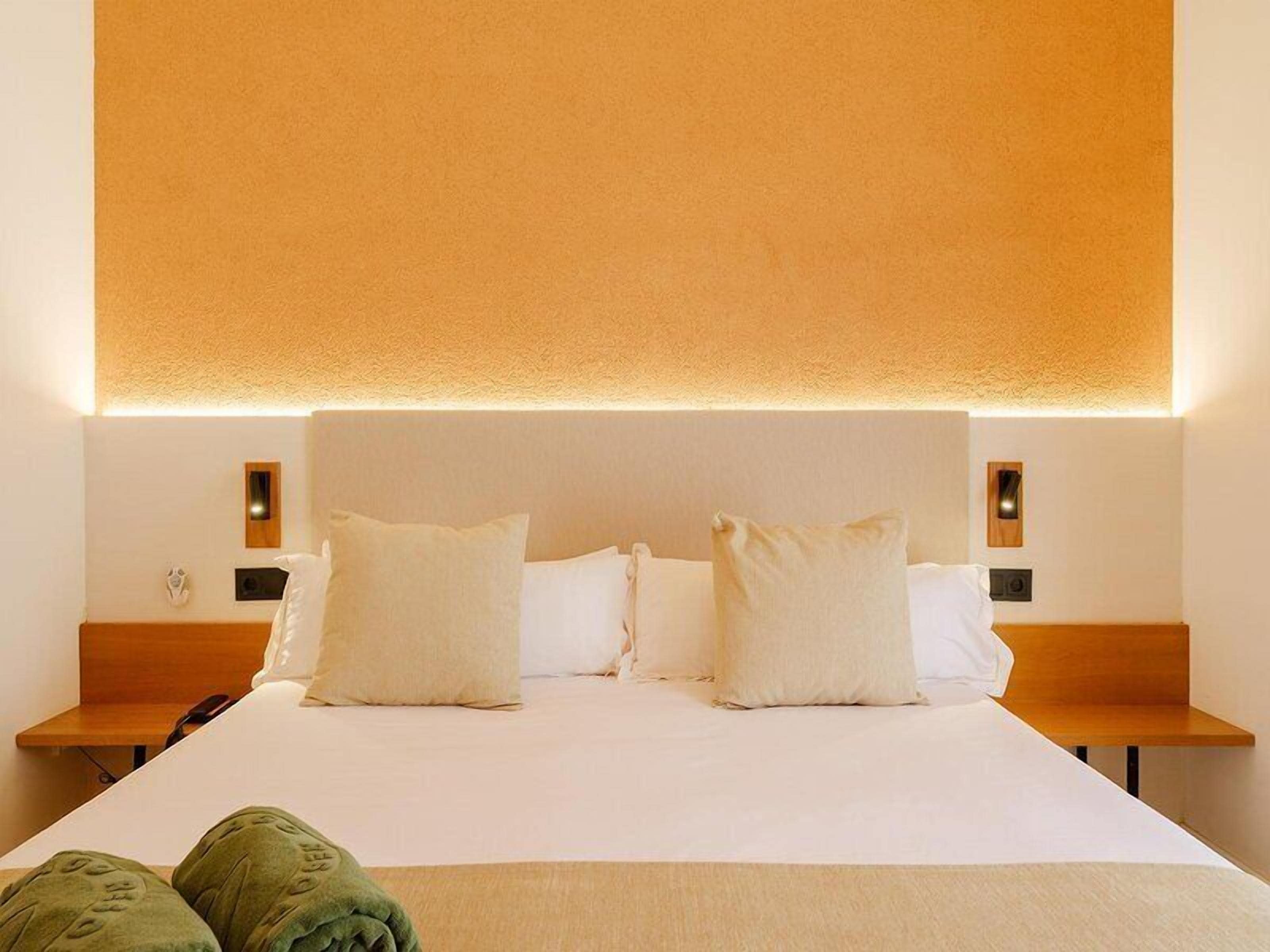 Quarto de hotel com uma cama grande, duas mesas de cabeceira e luminária fixada na parede.