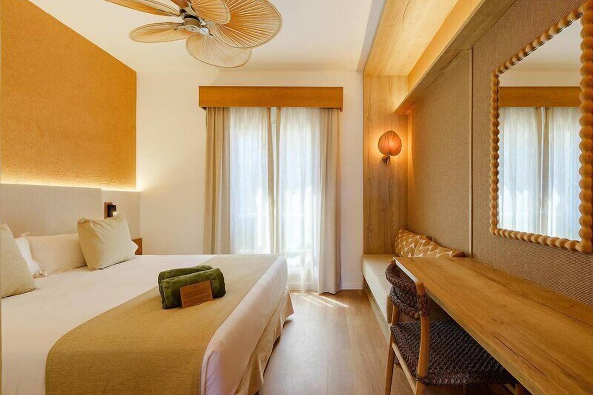 A hotel room with a bed, a desk, a chair, a large mirror, and a ceiling fan.