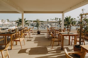 A covered outdoor dining area with wooden tables and chairs, overlooking a marina with boats and palm trees.