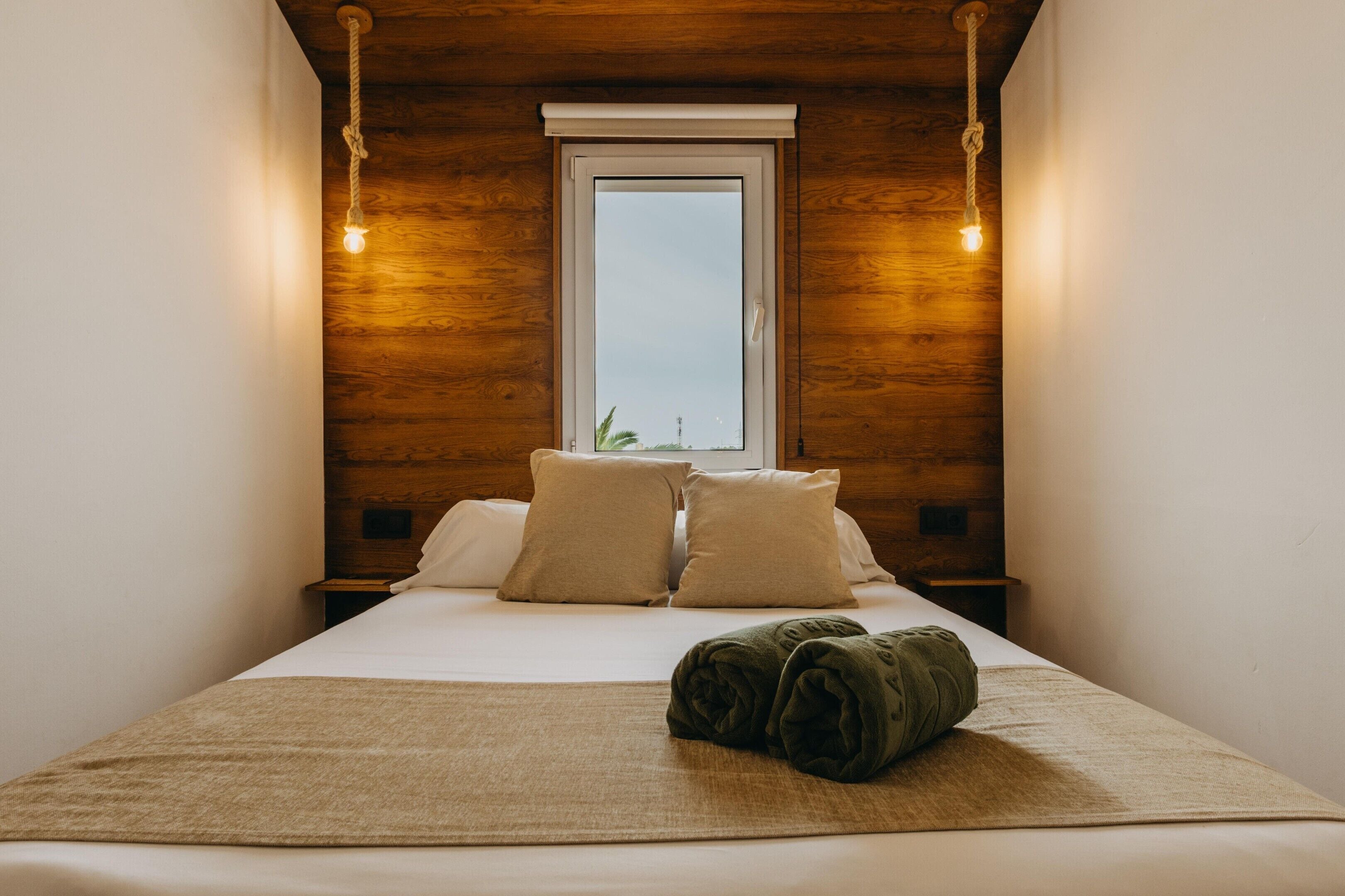 Una cama bien hecha con almohadas, una toalla enrollada y una ventana con vistas.