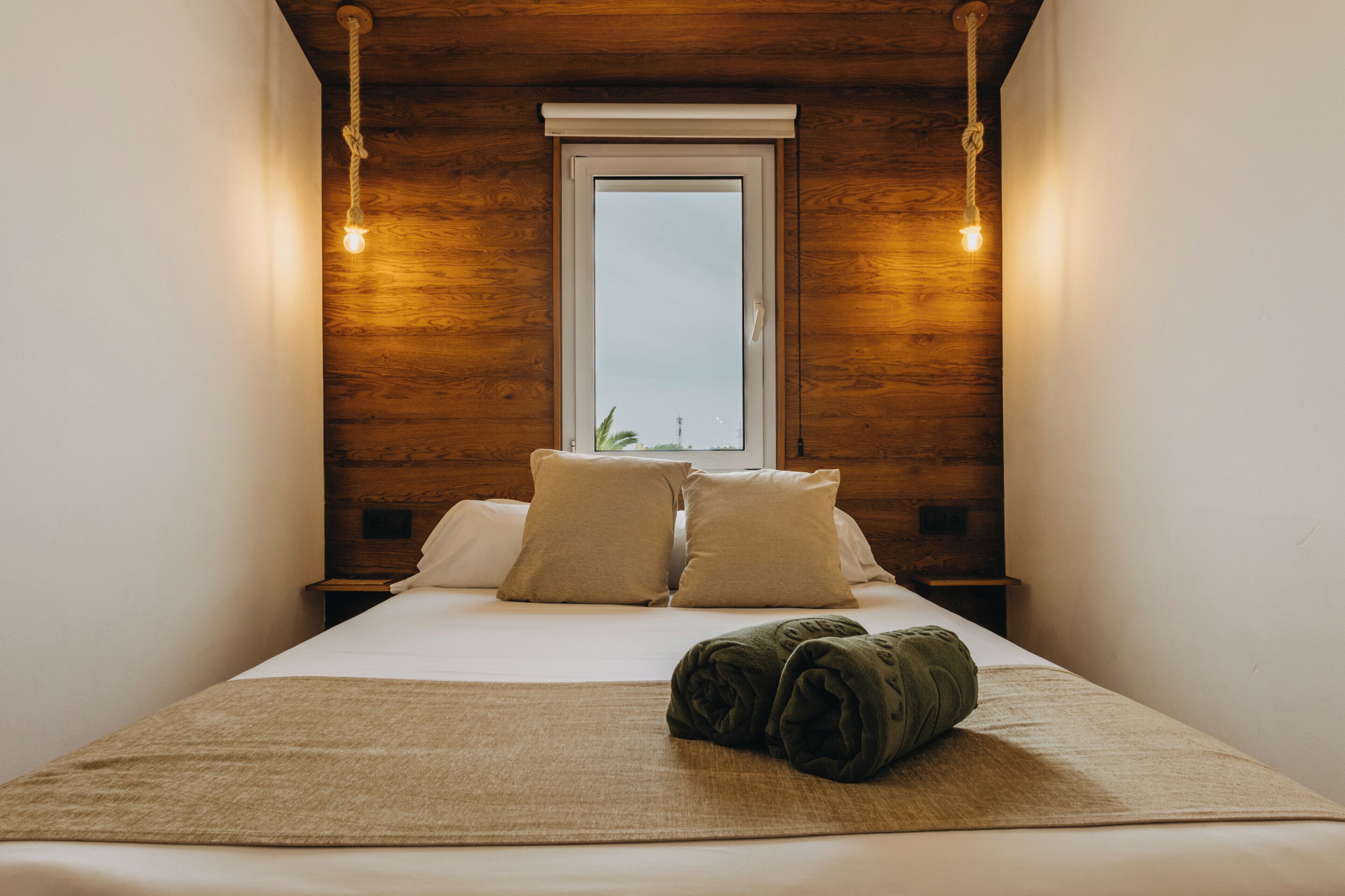 Ein Zimmer mit einem Bett, zwei Kissen, einem zusammengerollten Handtuch, einem Fenster und Holzwänden.
