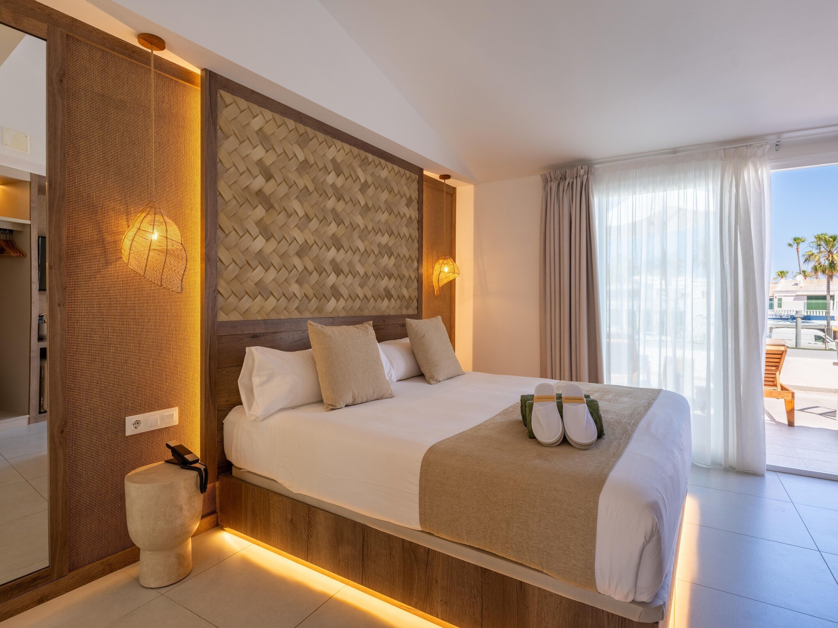 A hotel room with a large bed, a wooden headboard, a small bedside table, and a view of the outside.