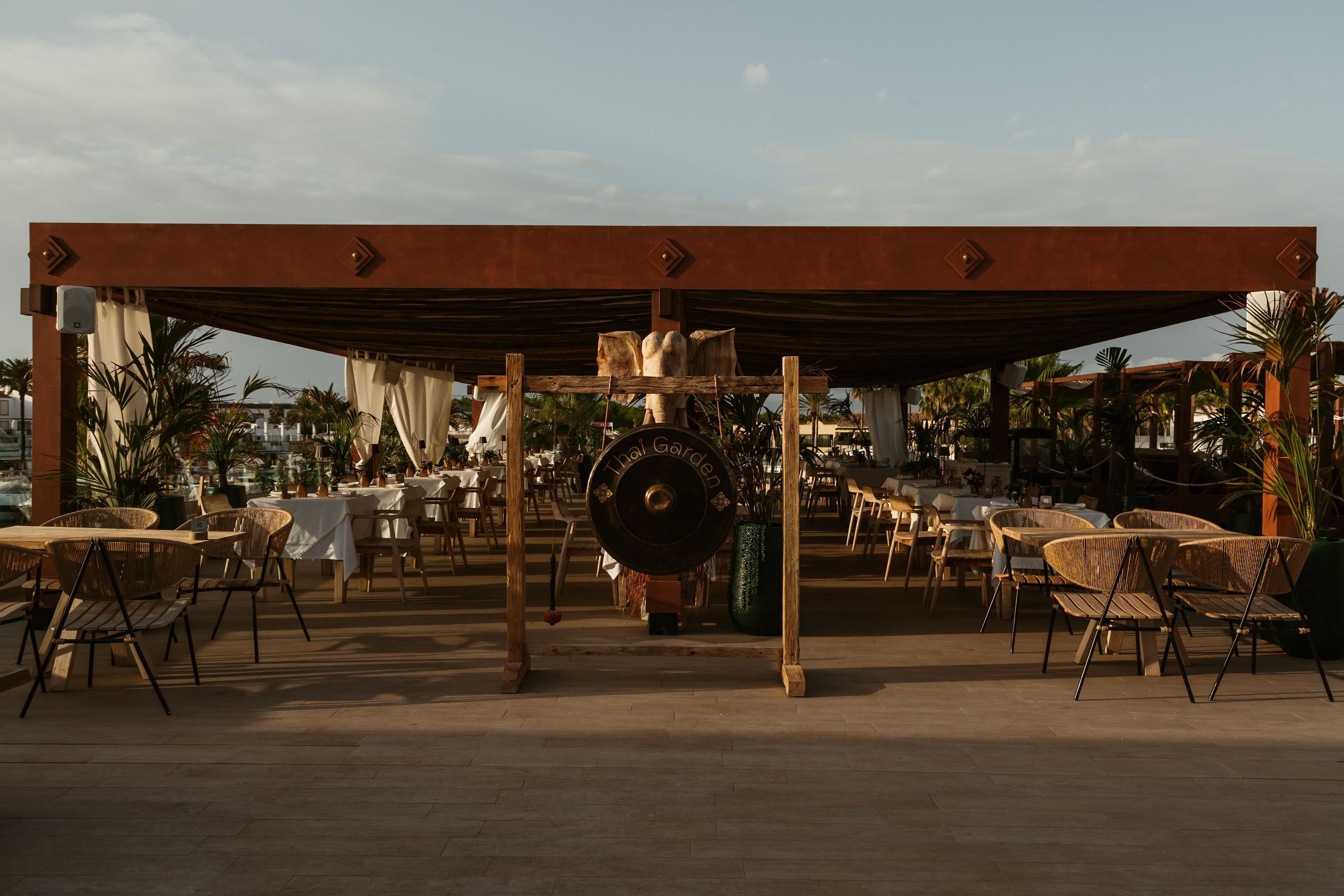 Un espace de restauration en plein air avec des chaises et des tables en osier, une pergola au-dessus et une sculpture métallique décorative.