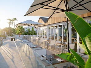 An outdoor dining area with tables and chairs, umbrellas, and a view of palm trees. - Lago Resort Menorca Casas del Lago - Adults Only (Ciutadella de Menorca)