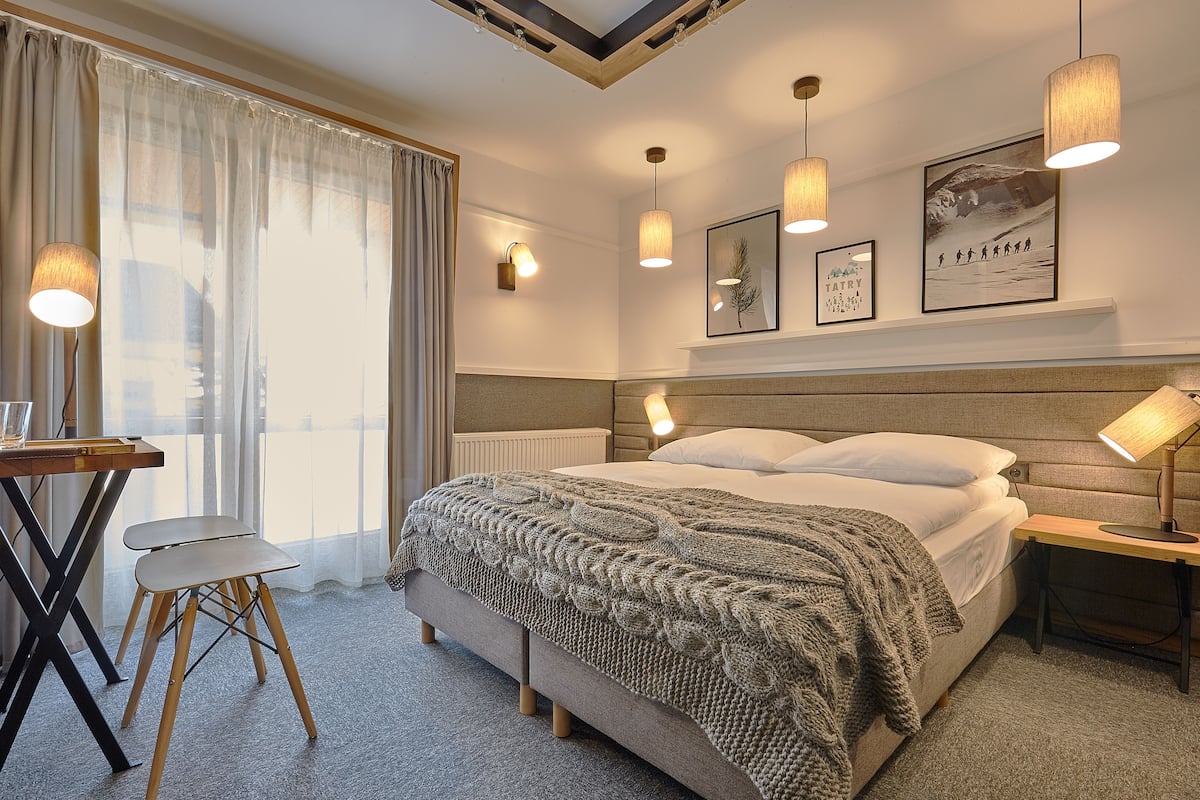 A neatly arranged hotel room with a bed, bedside tables, a desk with a chair, and a large window with curtains.