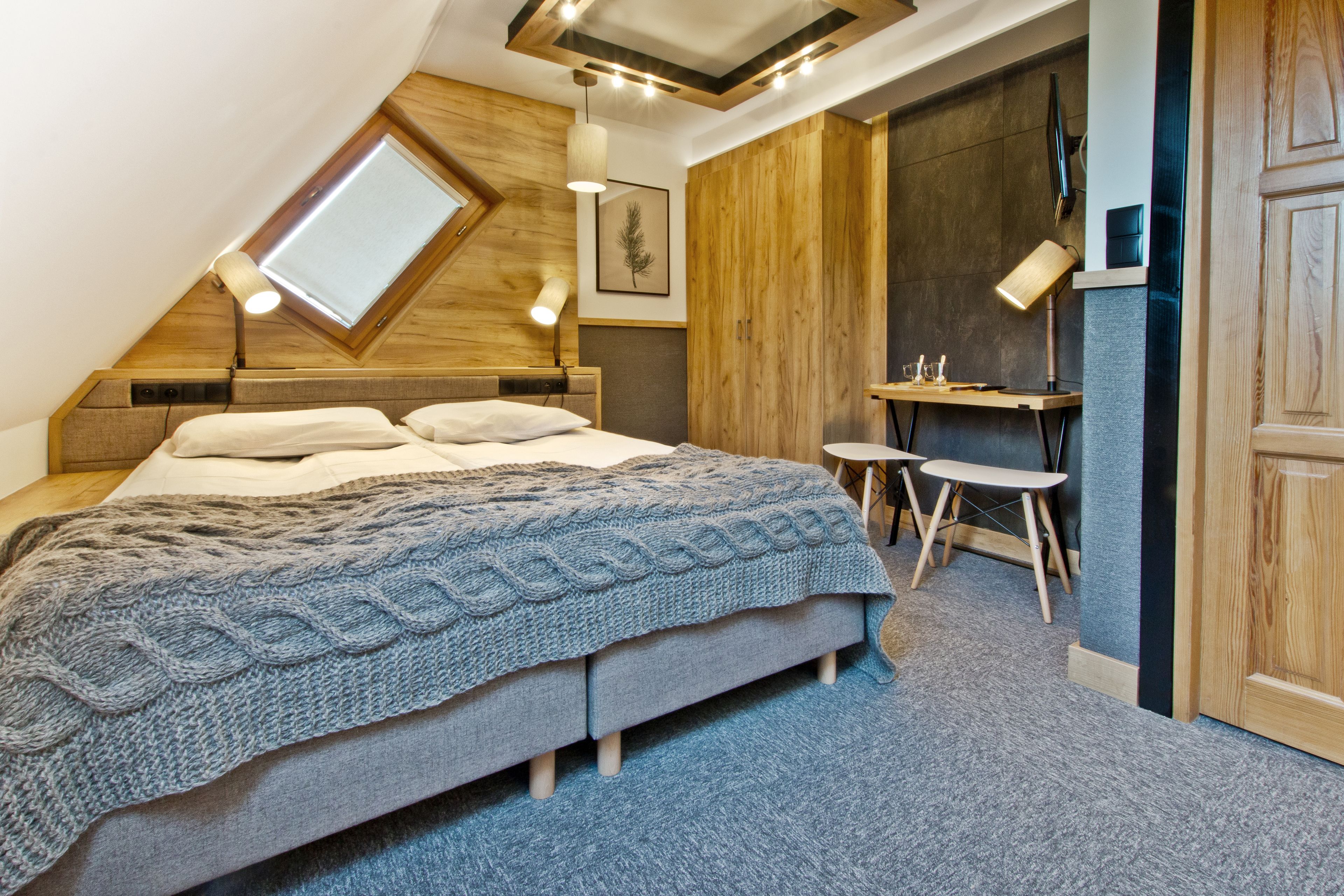 A bedroom with a sloped ceiling, a bed with a knitted blanket, a desk with two chairs, and a wooden wardrobe.