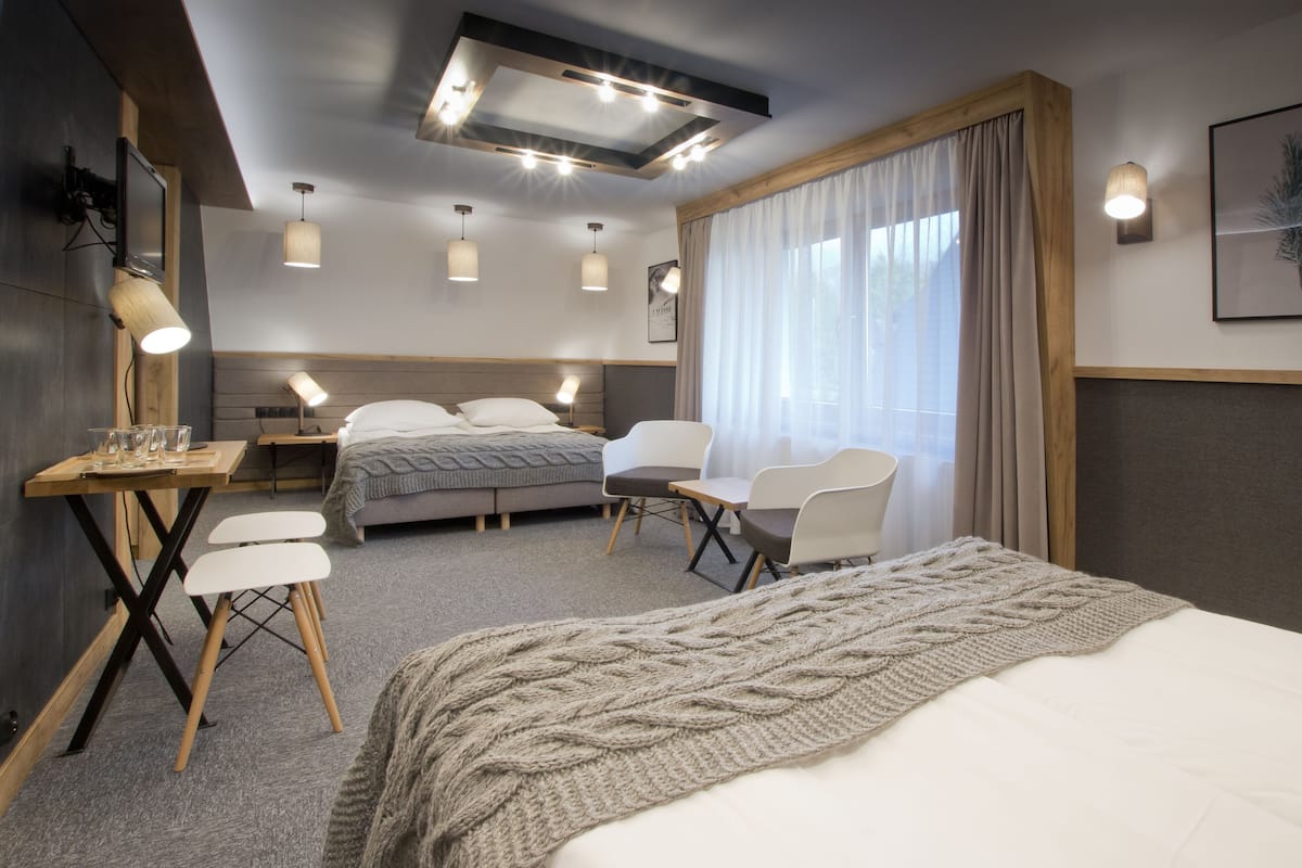A modern hotel room with a large bed, a desk with a chair, a small table, and a TV. There are two chairs and a window with curtains.