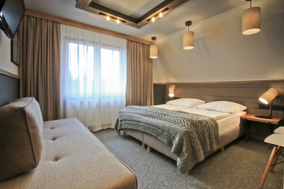 A modern hotel room with a large bed, a sofa, a desk, and a window with curtains.