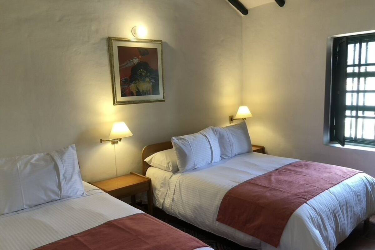 Une chambre avec deux lits, chacun recouvert d’une literie blanche et d’une couverture rouge. On y trouve des lampes de chevet, un tableau encadré au mur et une fenêtre avec des stores.