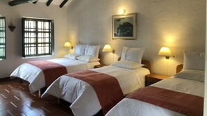 A room with three beds, each with white bedding and a brown throw. There are wall-mounted lamps on either side of the beds. A window with a grid pattern is visible on the left wall. - Hotel Hospedería San Carlos Villa de Leyva (Villa de Leyva)