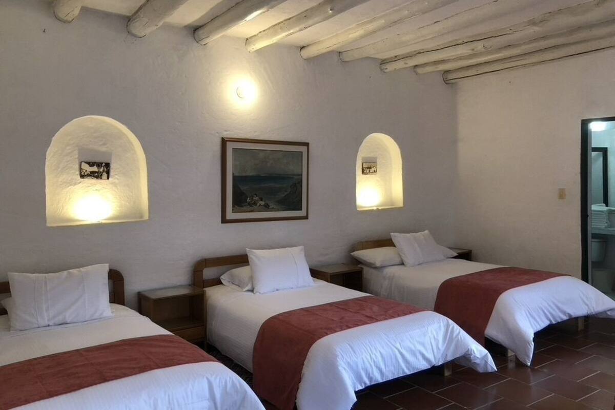 Une chambre d’hôtel avec trois lits, chacun agrémenté de draps blancs et de touches de couleur rouge. Des lampes murales et un tableau encadré sont fixés au mur au-dessus du lit central.