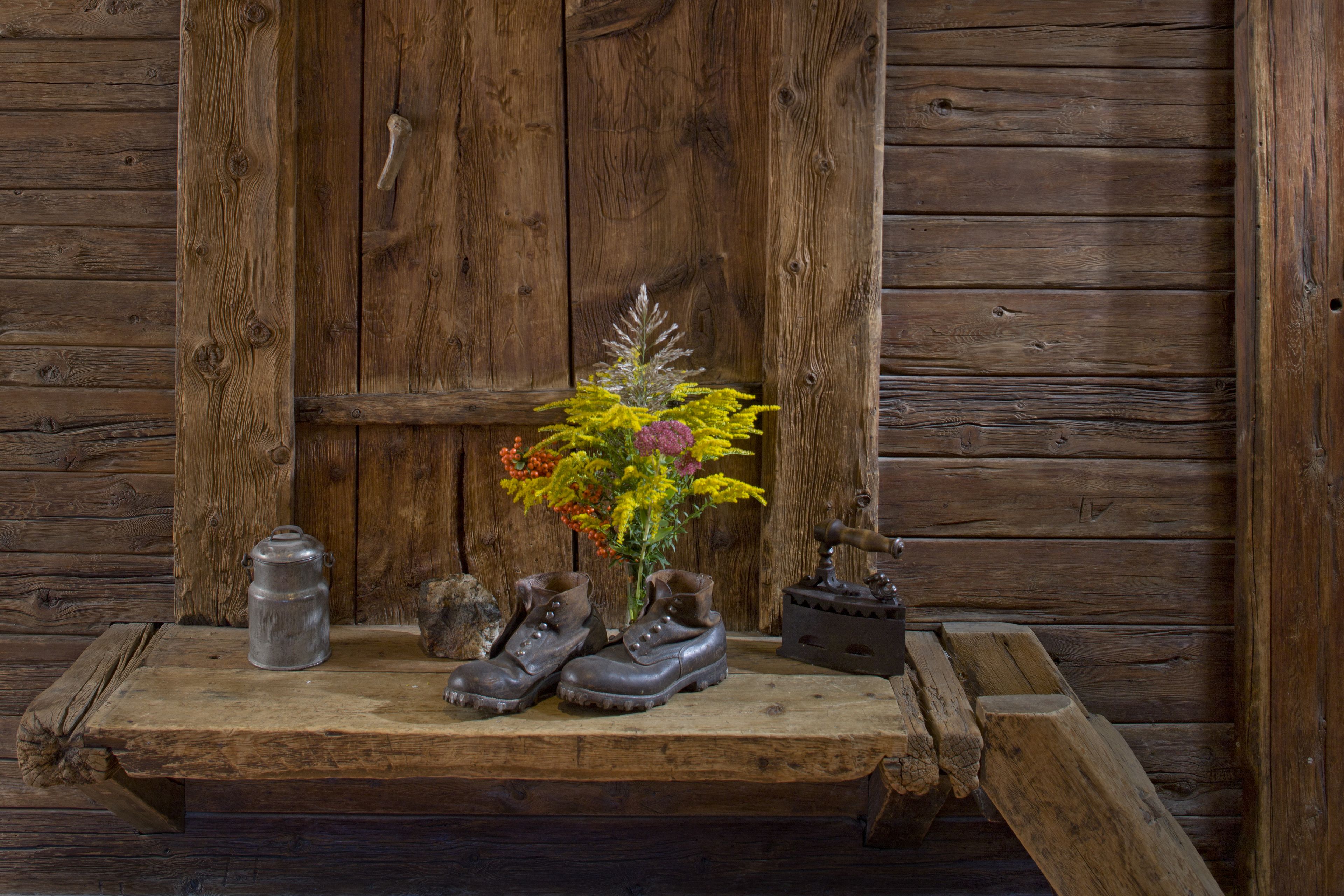 A rustic wooden bench with a pair of boots, a metal container, and a small vase with flowers against a wooden wall.