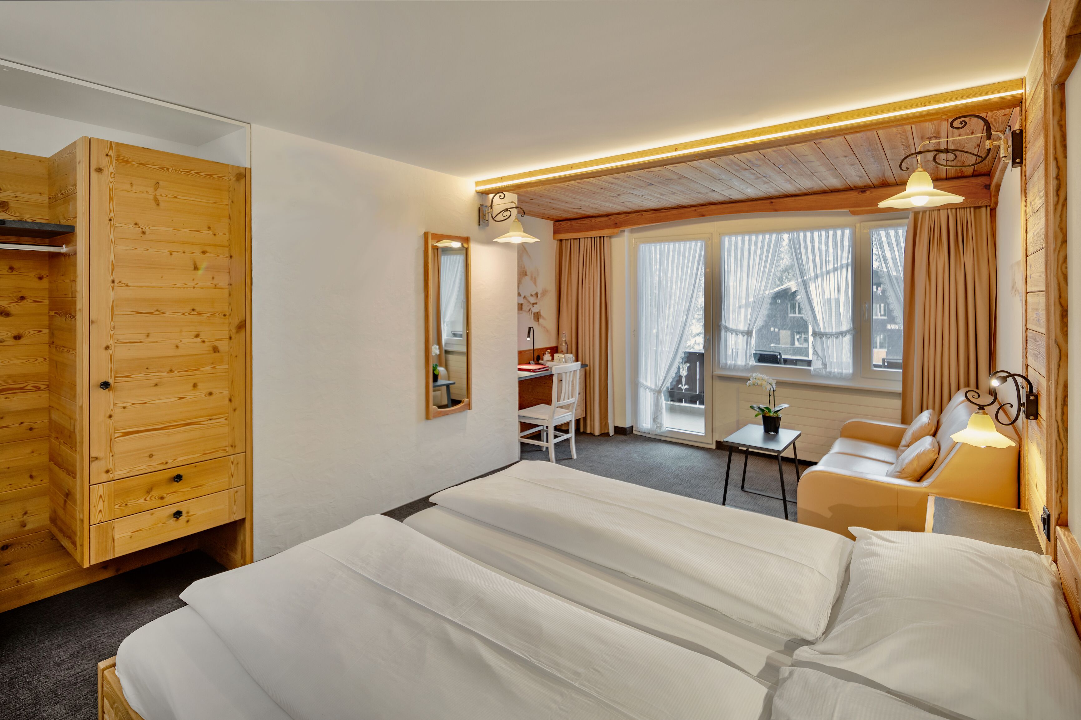 Une chambre d’hôtel avec une armoire en bois, un lit avec du linge de maison blanc, un petit bureau avec une chaise, un canapé et une fenêtre avec des rideaux.