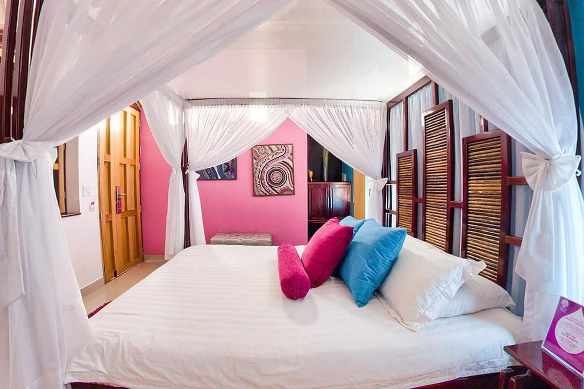 A bedroom with a bed, white curtains, a pink wall, and a wooden headboard.