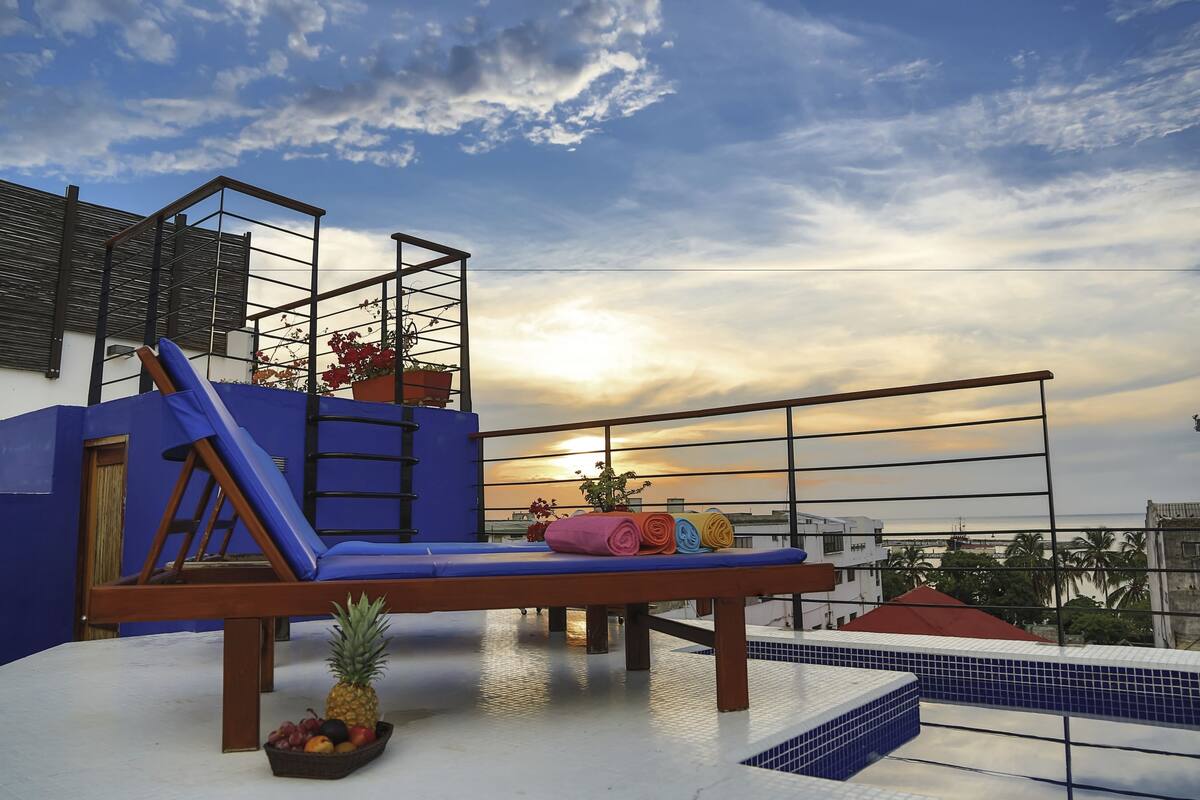 A rooftop terrace with a wooden lounge chair, a small pool, and a view of the sunset over the cityscape.