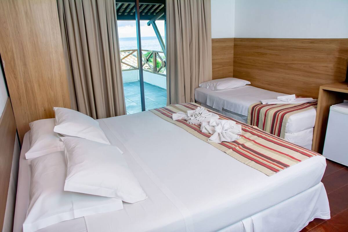 Une chambre d’hôtel avec un grand lit, deux petits lits, une tête de lit en bois et une vue sur la mer grâce à de grandes fenêtres.