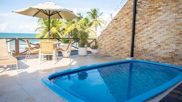 Una piscina con borde de baldosas, rodeada de sillas de mimbre y una mesa con sombrilla.
