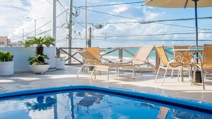 Una piscina con sillas de descanso y sombrillas, rodeada de plantas en macetas y un cielo despejado.
