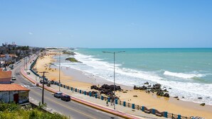 Una carretera costera con playa a la derecha, cielo despejado y olas rompiendo en la orilla.