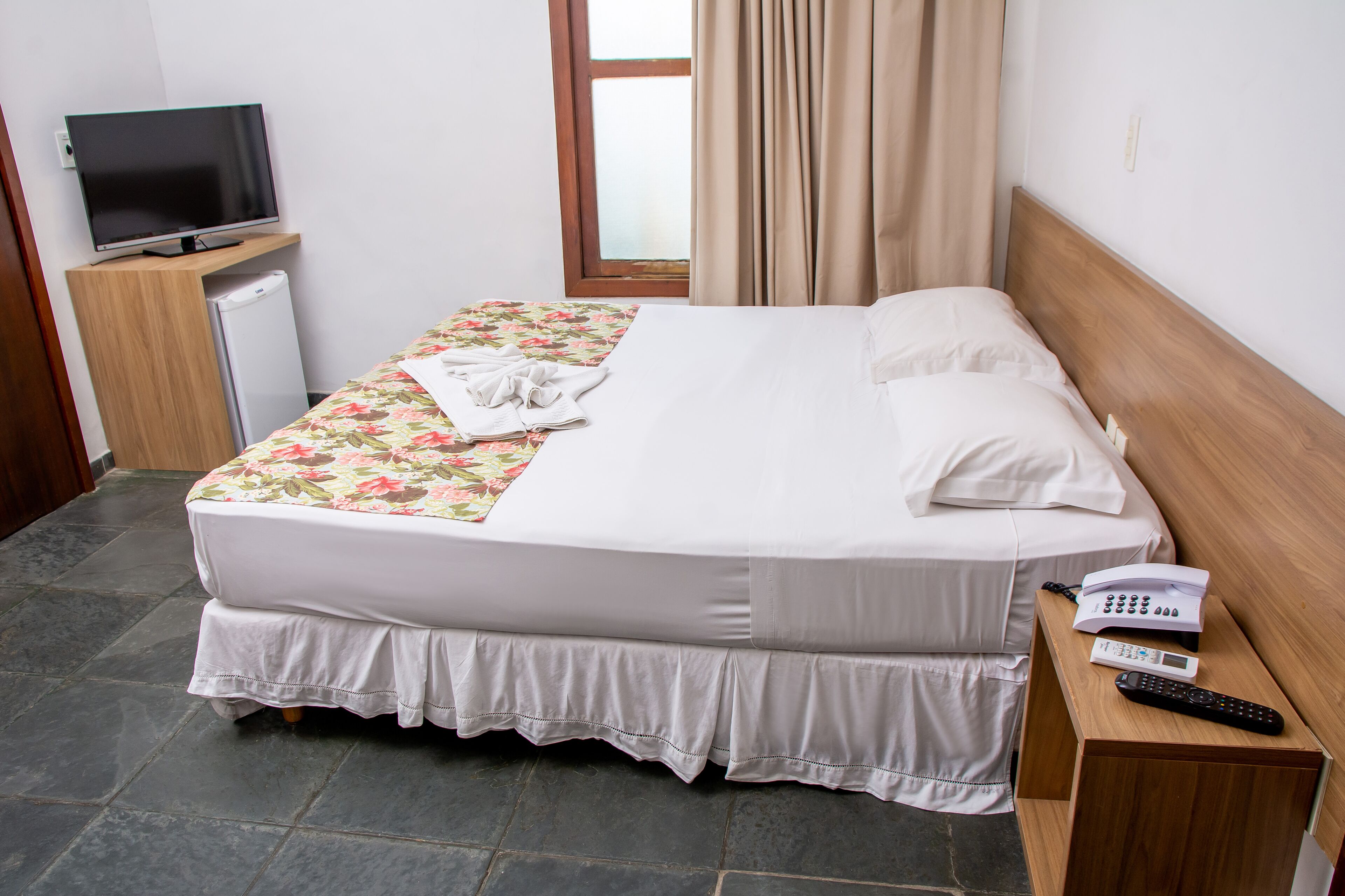 Un lit bien fait, recouvert d’une couvre-lit à motifs floraux, une table de chevet avec un téléphone et une télécommande, et un téléviseur posé sur un meuble à côté du lit.