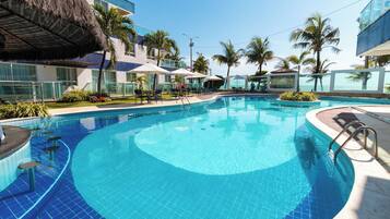 A swimming pool with lounge chairs and umbrellas, surrounded by palm trees and a building with a thatched roof.