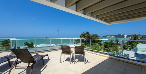 A balcony with two wicker chairs and a table, overlooking the ocean and a mountainous landscape.