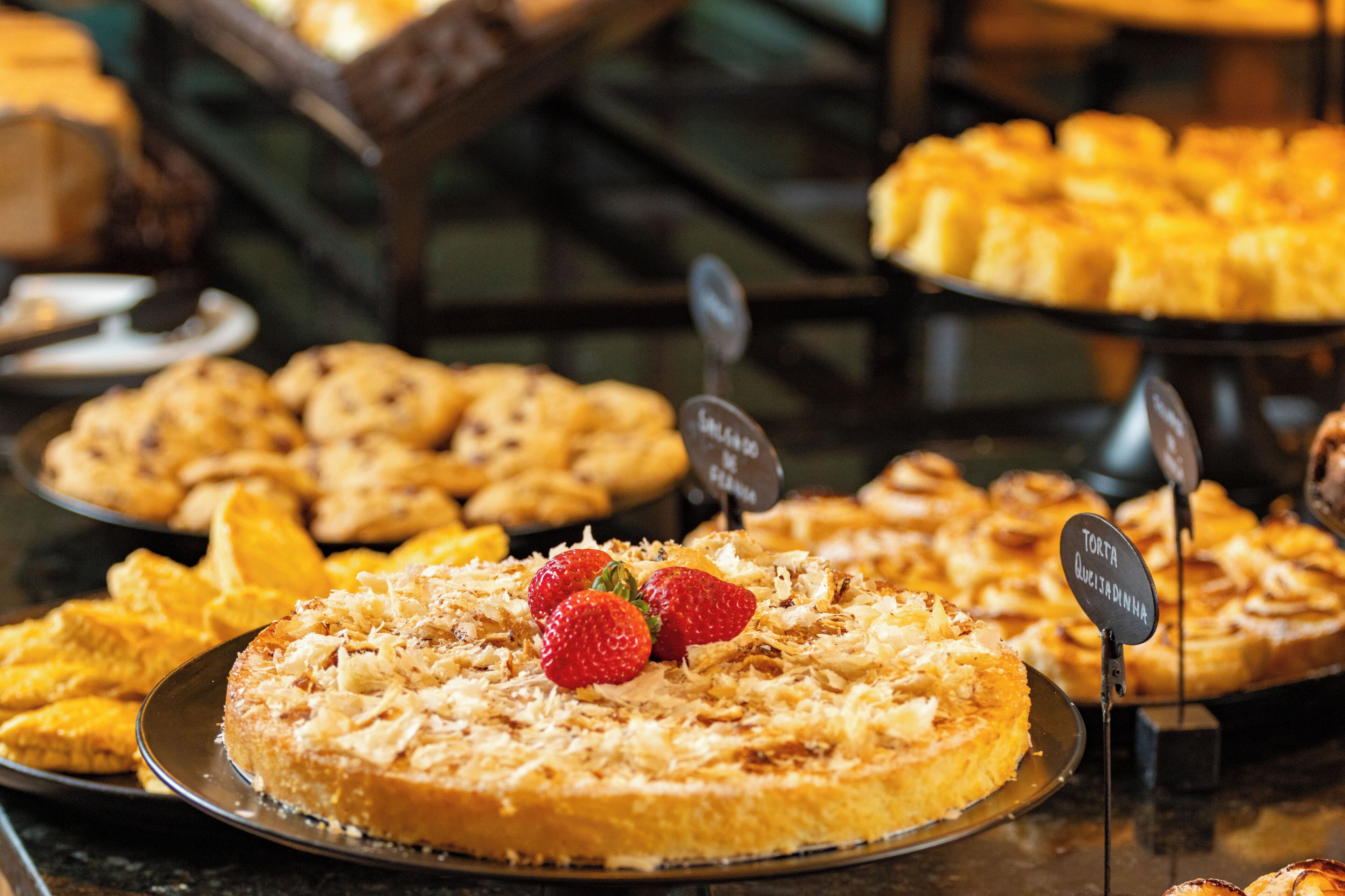 Exposição de diversos produtos de pastelaria, incluindo um bolo com morangos, uma placa com a inscrição 'TORTA QUETADINA' e outras iguarias.