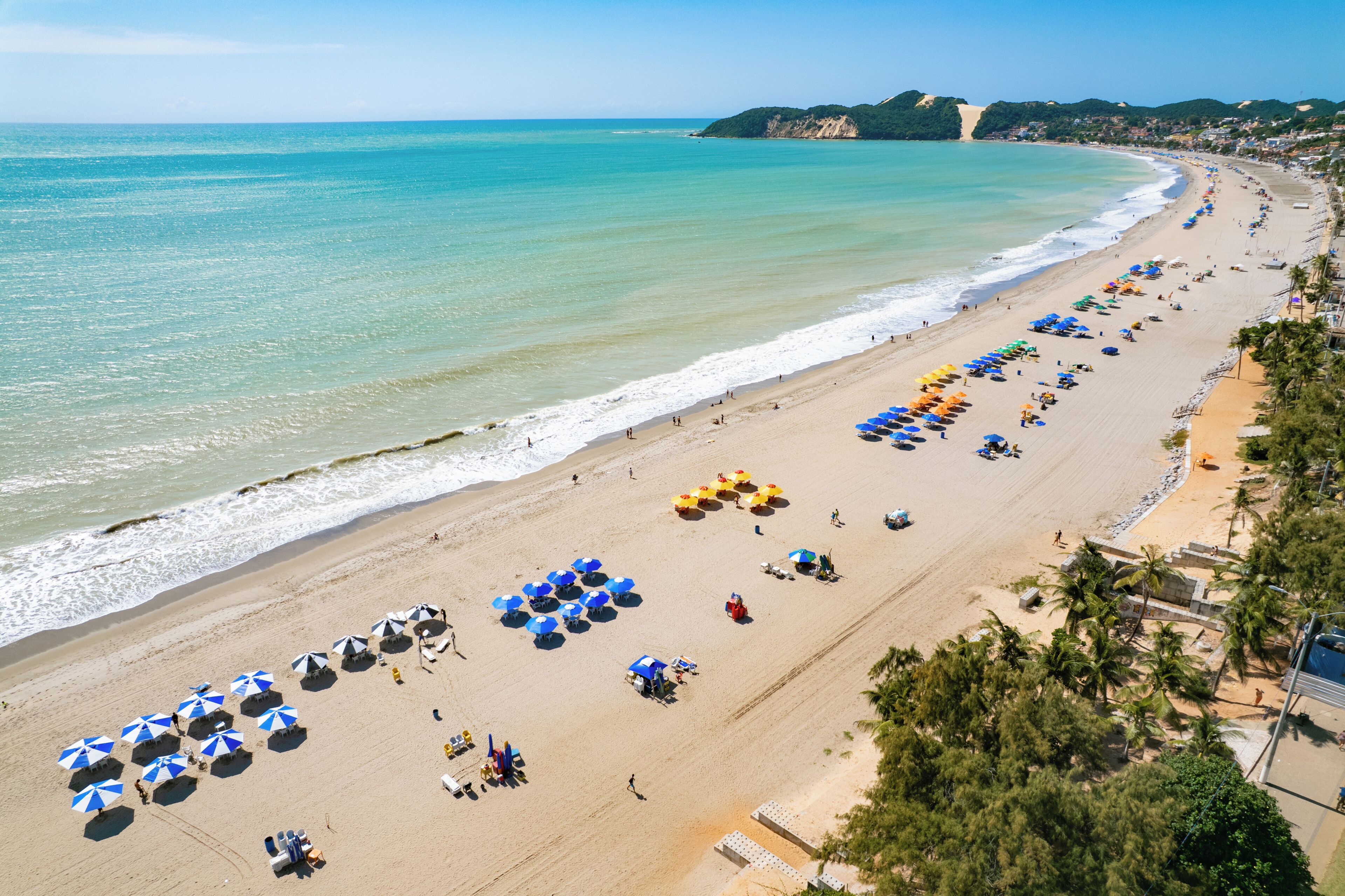A beach with rows of umbrellas and sun loungers, a clear sky, and a coastline with buildings in the distance.