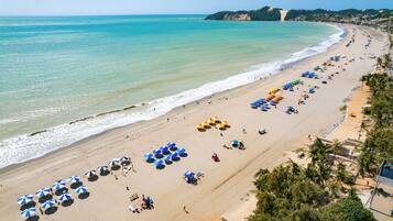 A beach with rows of umbrellas and sun loungers, a clear sky, and a coastline with buildings in the distance.