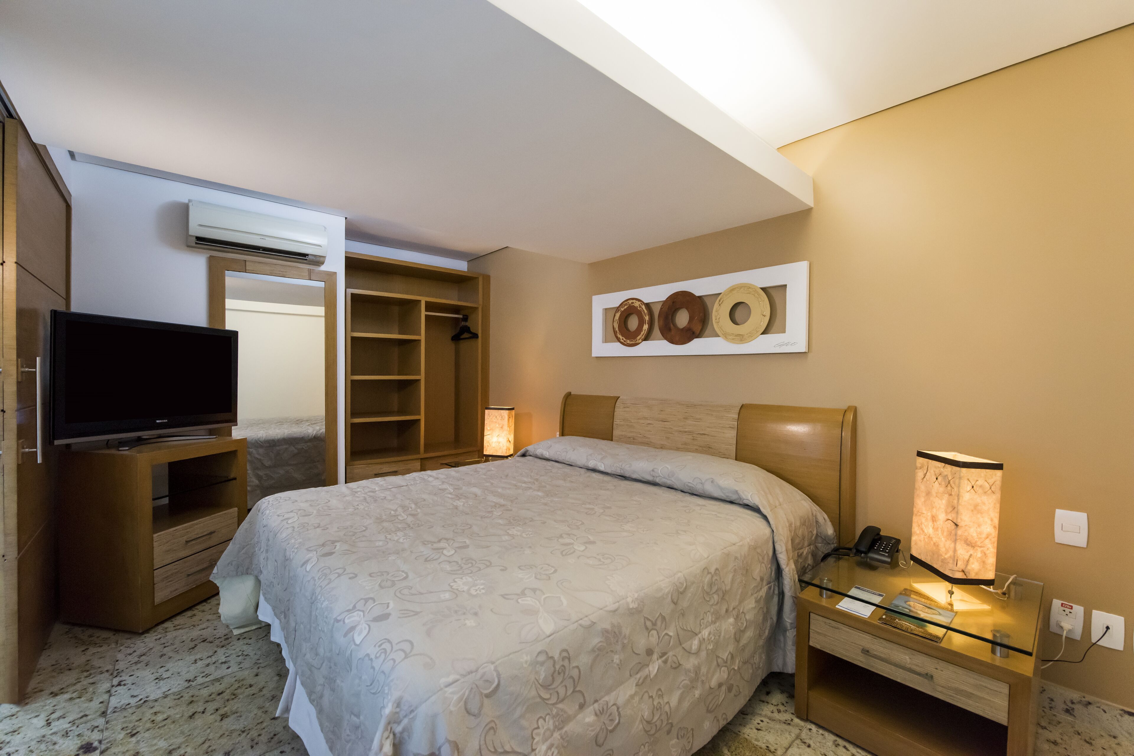 Une chambre d’hôtel comprenant un lit, une table de chevet avec une lampe, une télévision, une armoire et une œuvre d’art accrochée au mur.
