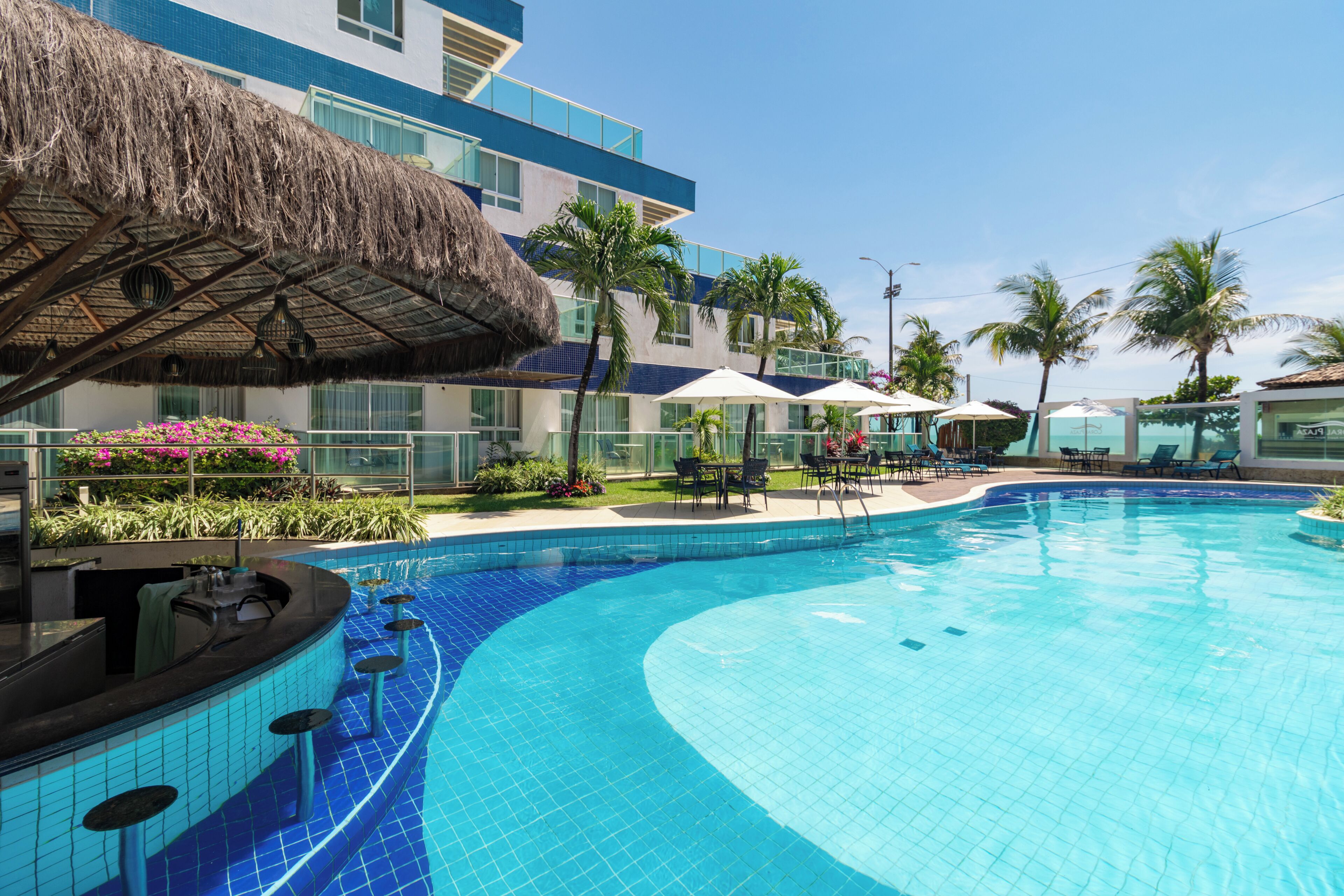 Ein Pool mit geschwungener Kante, umgeben von Liegestühlen und Sonnenschirmen, mit einem Gebäude mit blau-weißer Fassade und einem Reetdach.