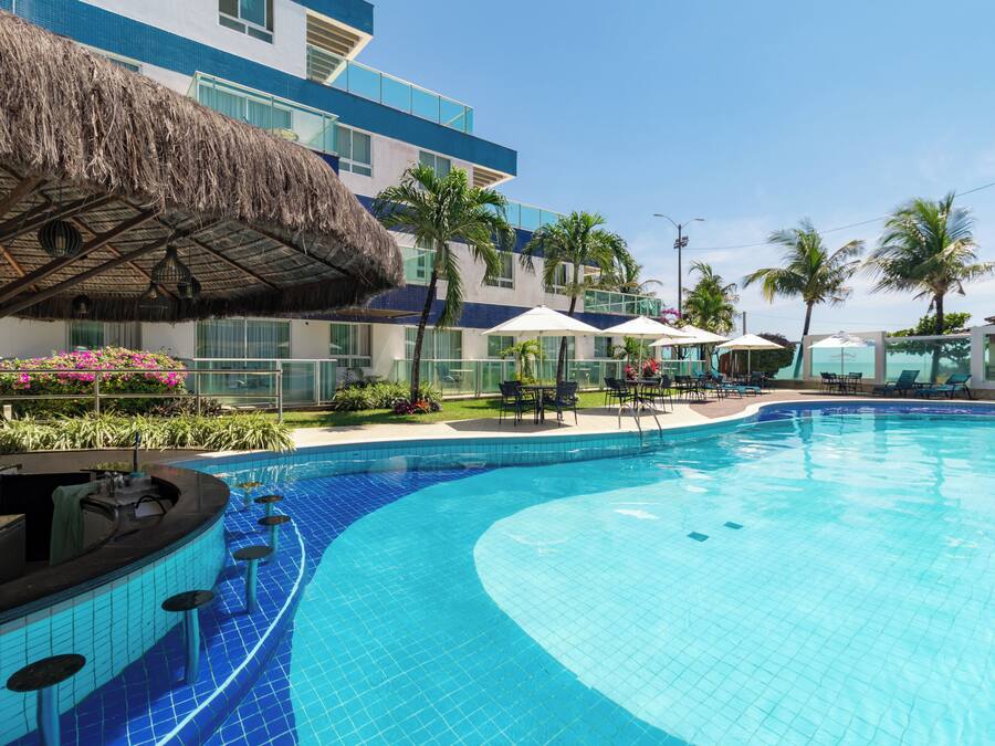 Ein Pool mit geschwungener Kante, umgeben von Liegestühlen und Sonnenschirmen, mit einem Gebäude mit blau-weißer Fassade und einem Reetdach.